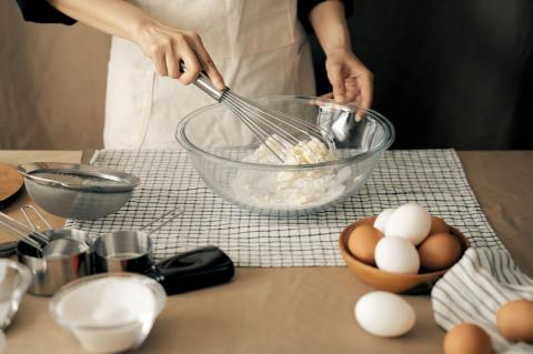 hands of woman wearing an apron, mixing ingredients in a glass bowl with eggs sitting in another bowl on a table with measuring cups and baking utensils